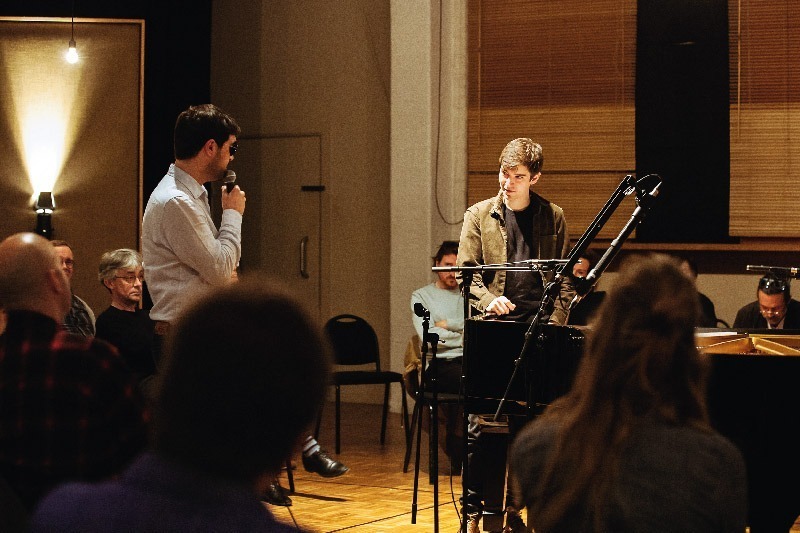 Karl Meesters, holding a microphone, adresses Bram De Looze, leaning on the grand piano