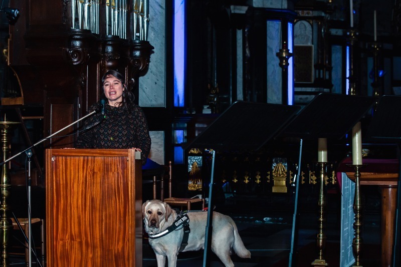 Une dame accompagnée d'un chien d'assistance s'adresse à l'auditoire depuis l'arrière du pupitre.