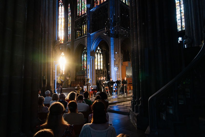Vooraan in de kerk staan de 7 zangers van Dionysos Now. Achter hen schijnt het licht van de ondergaande zon door de glas-in-loodramen.