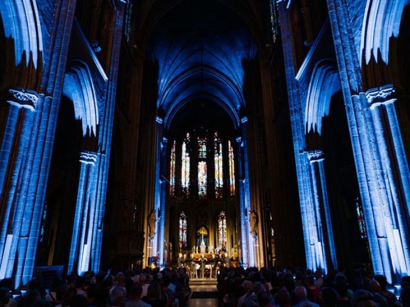 Vue vers l'autel où se tiennent les chanteurs de Dionysos Now ! À l'intérieur de l'église, il fait sombre, la structure étant éclairée par des lumières bleues.