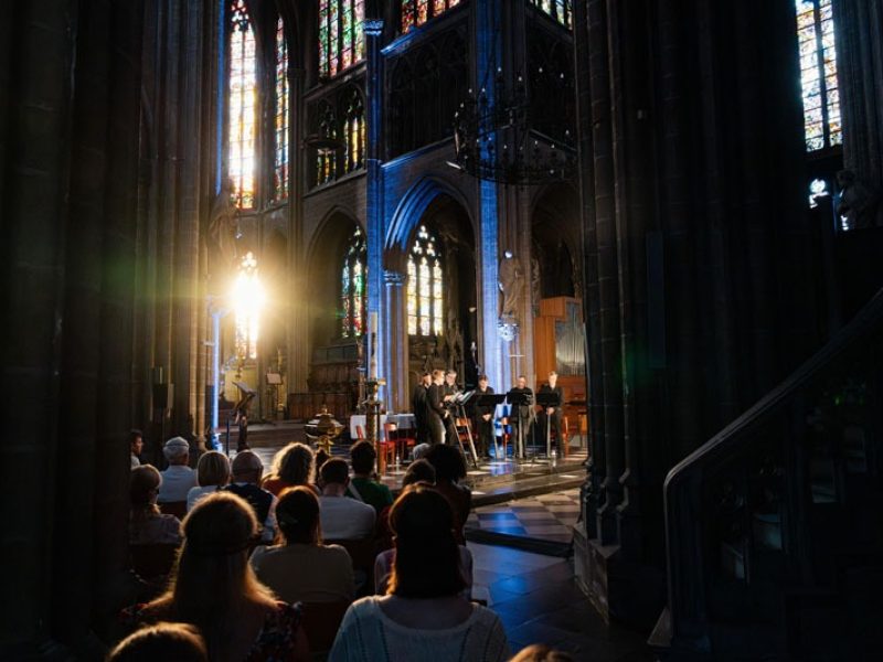 Sur le devant de l'église se trouvent les sept chanteurs de Dionysos Now! Derrière eux, la lumière du soleil couchant traverse les vitraux.